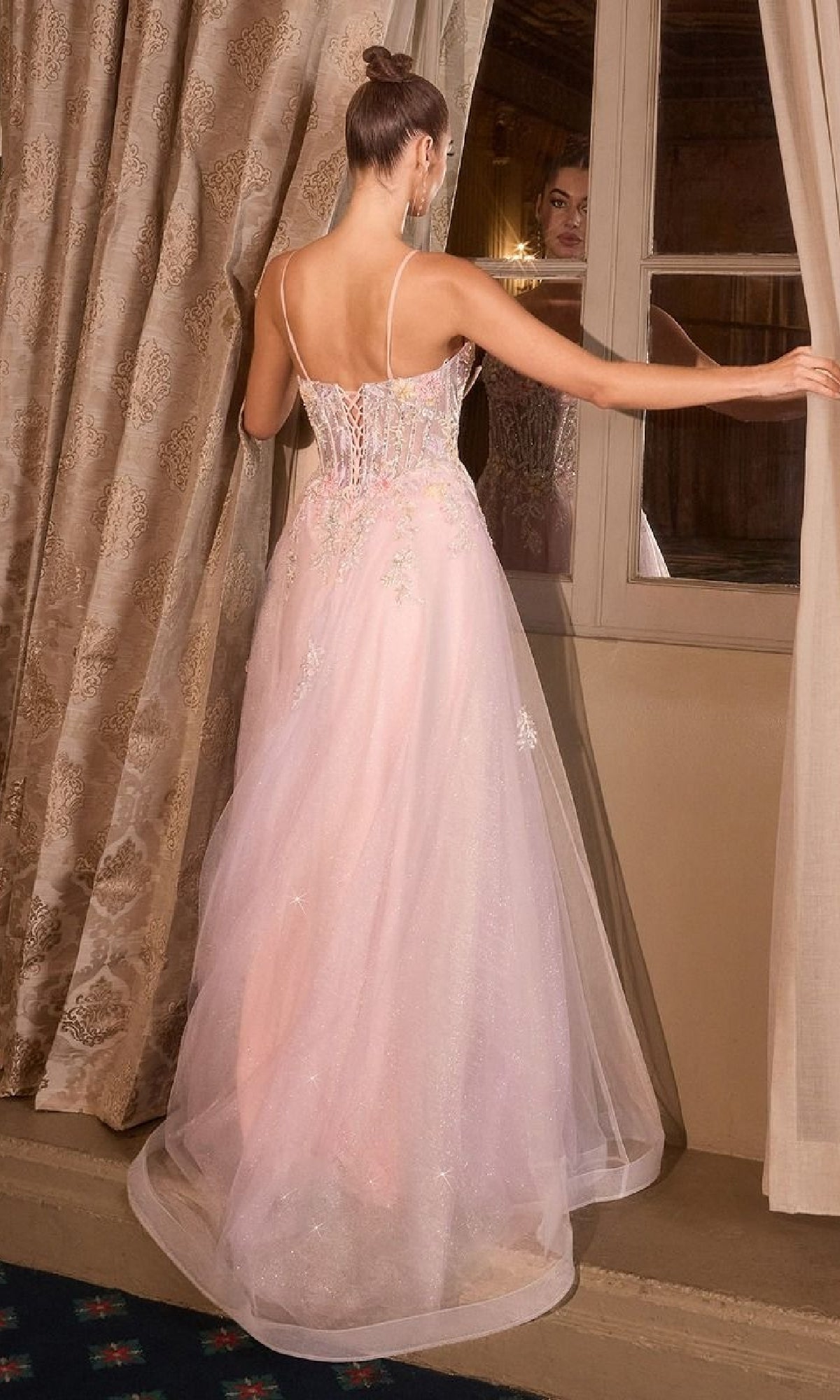 A woman wearing the Classic Long Pink Prom Dress: Andrea and Leo A1477, featuring thin straps and a corset back, stands by a window with her back to the camera, gazing at her reflection.